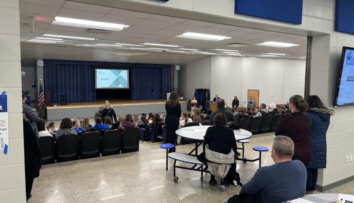 View looking in to the Butler Middle School auditorium, where the seats are full and there are people standing behind.