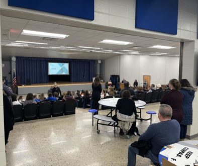 View looking in to the Butler Middle School auditorium, where the seats are full and there are people standing behind.